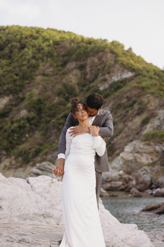 Groom giving a cheek kiss to the bride and hugging her from behind after their private elopement on a cliffside in Greece.